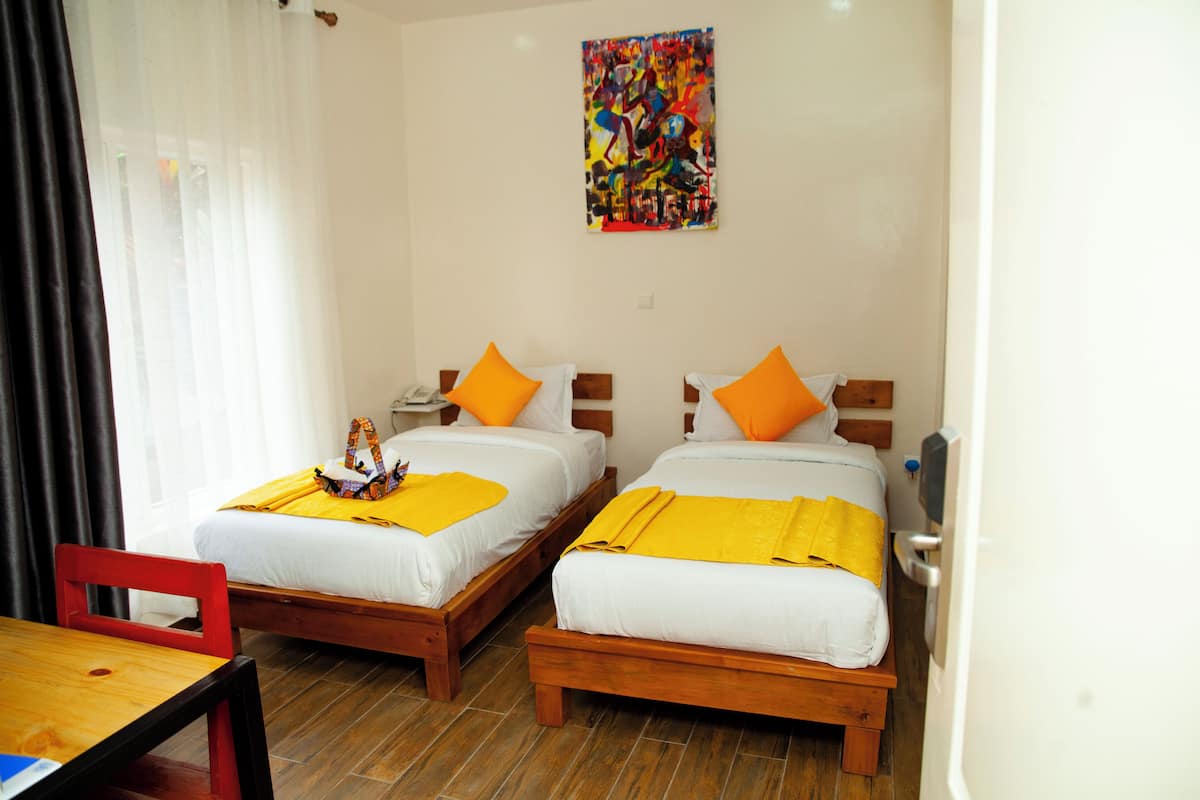 A hotel room with two wooden beds, each with white bedding and orange pillows. There is a wooden table with a chair, a colorful abstract painting on the wall, and a window with sheer curtains.