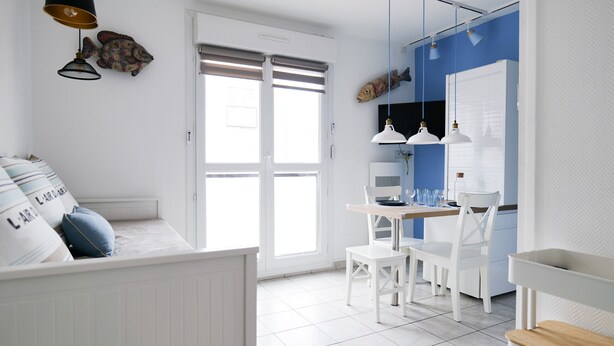 A modern kitchen with white cabinetry, a dining table, and chairs. There is a large window with blinds, and a white bench with pillows in the foreground.