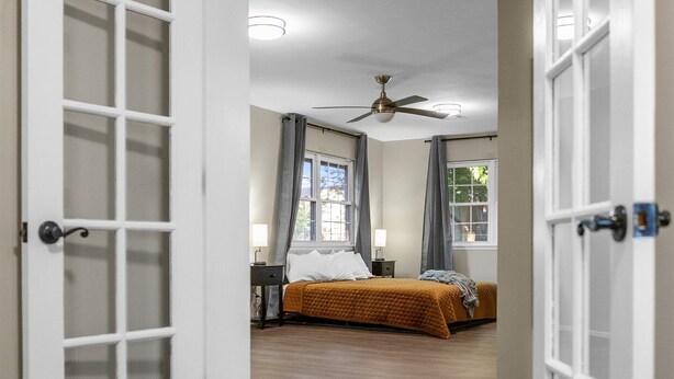Ein Flur, der zu einem Schlafzimmer mit Bett, Deckenventilator und einem mit Vorhängen drapierten Fenster führt.