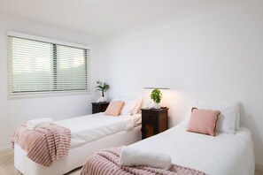 Une chambre avec deux lits, une table de chevet, une plante en pot et une fenêtre avec des stores.