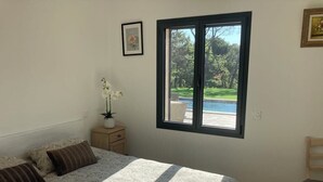 Un dormitori amb un llit gran, una finestra amb vista a una piscina i vegetació, una taula petita amb un ram de flors, i una cadira.