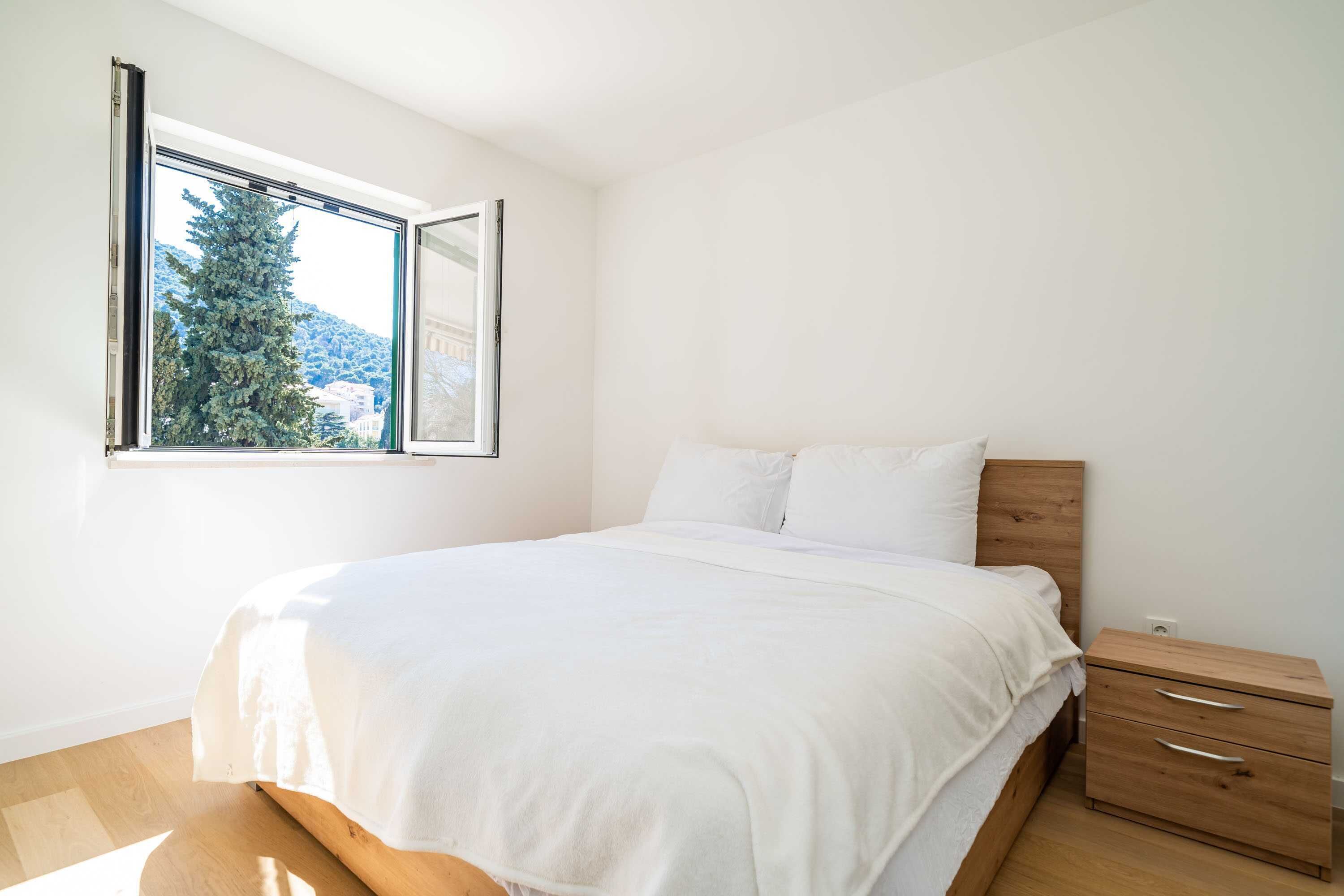 Ein Schlafzimmer mit Bett, Nachttisch, Fenster mit Blick auf Baum und Berge sowie einer weißen Wand.