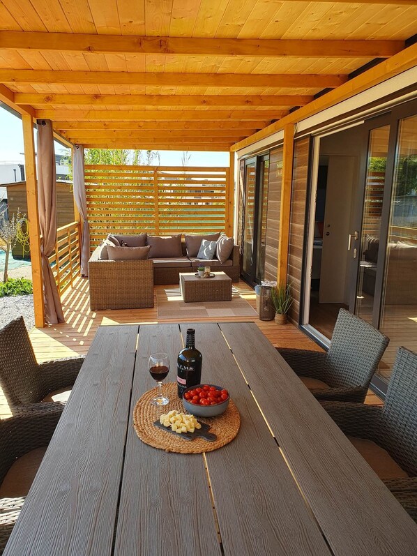 Un patio cubierto con una mesa de madera, mobiliario de mimbre y un ventanal de vidrio que da a una zona de piscina.