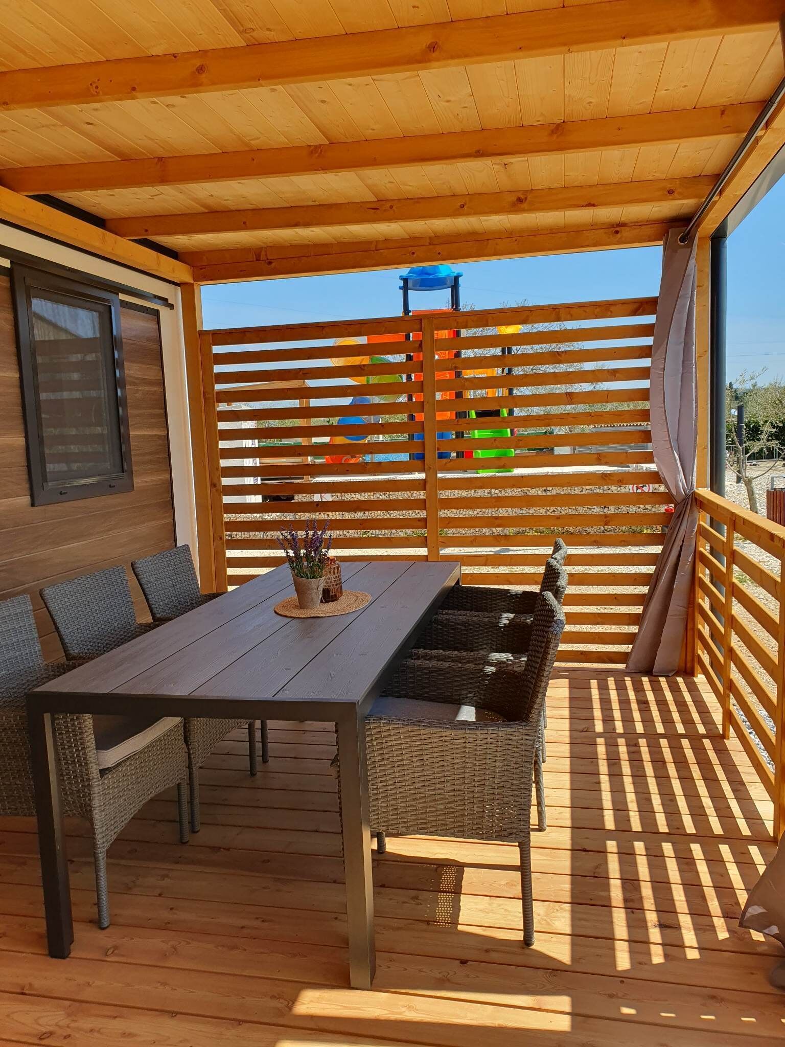 A covered outdoor seating area with a table and wicker chairs, a wooden deck, and a view of a playground in the background.