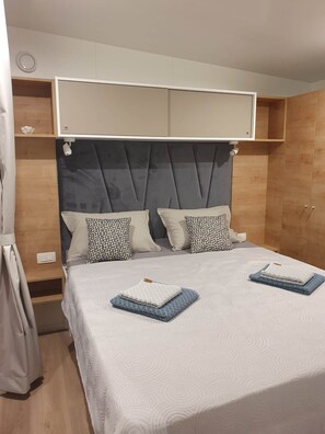 Una cama doble con cabecera tapizada en gris, dos mesitas de noche y un espejo grande sobre la cama.