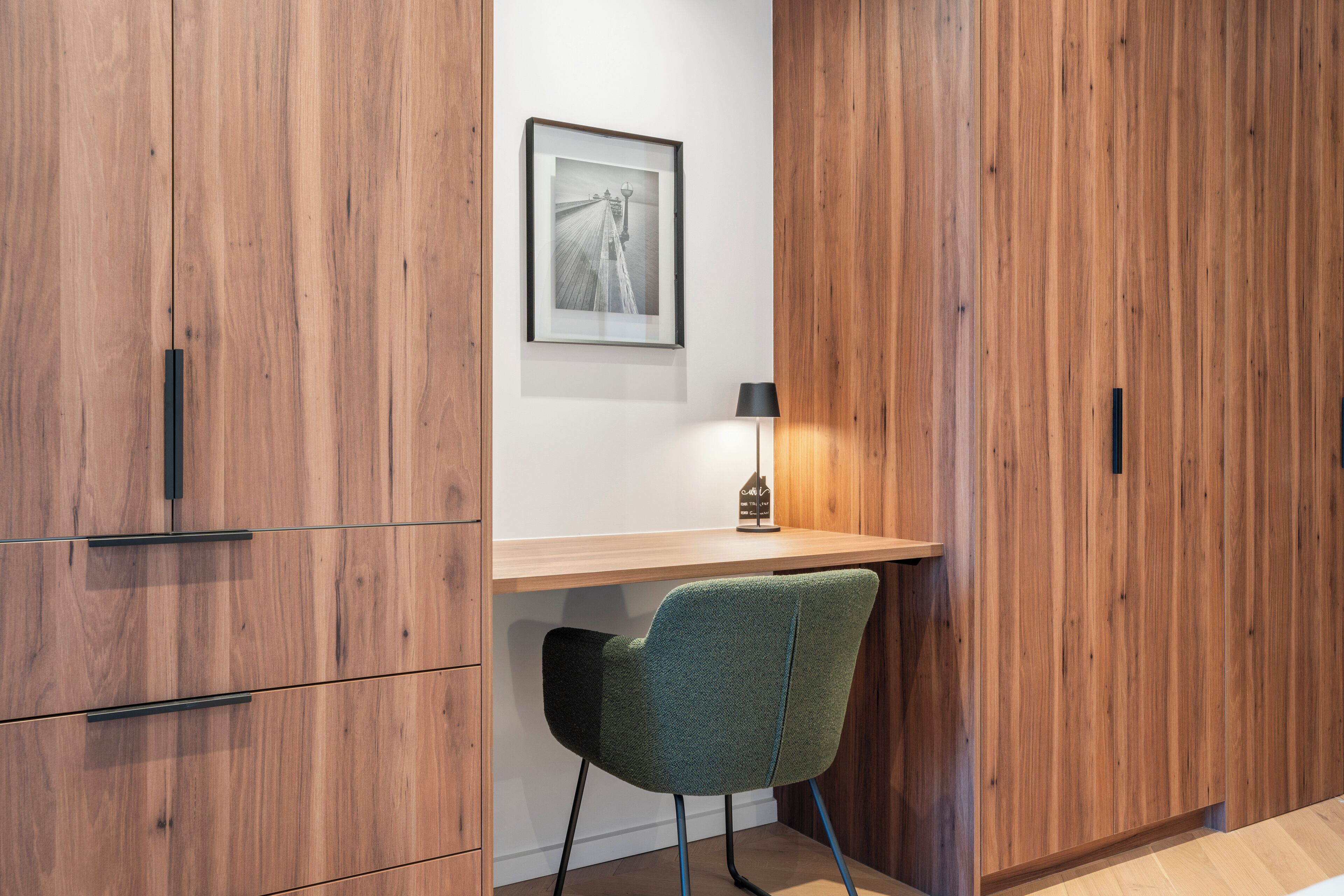 A wooden wardrobe with a framed picture on the wall, a desk with a lamp, and a green chair.