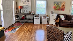 A cozy living room with a wooden floor, a brown leather sofa, a wicker chair, a white stove, a small white cabinet, and a colorful rug. - Family Hideaway ,family friendly and pet friendly. (Virginia Beach)