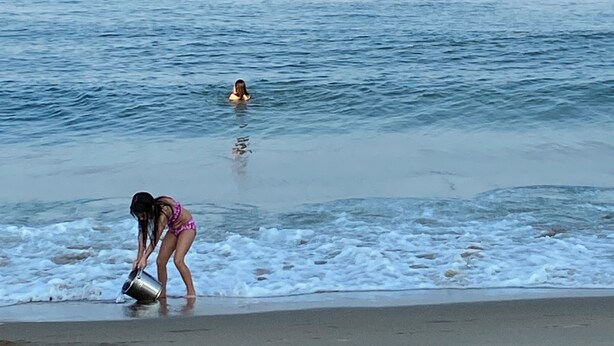 A beach scene with a person in a bikini bending over near the water's edge, another person swimming in the distance, and a boat visible on the horizon. - Family Hideaway ,family friendly and pet friendly. (Virginia Beach)