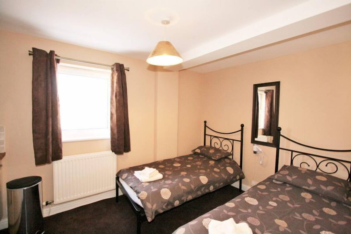 A small bedroom with two single beds, a radiator, a window with curtains, a hanging light fixture, and a mirror on the wall.