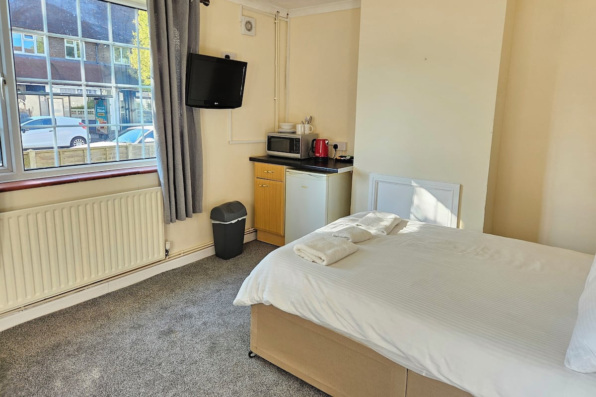 A hotel room with a bed, a small kitchenette, a TV, a window with curtains, and a radiator.