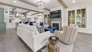 A spacious living room with a white sofa, patterned armchair, and a blue coffee table. A television is mounted above a fireplace, and there is a kitchen in the background.