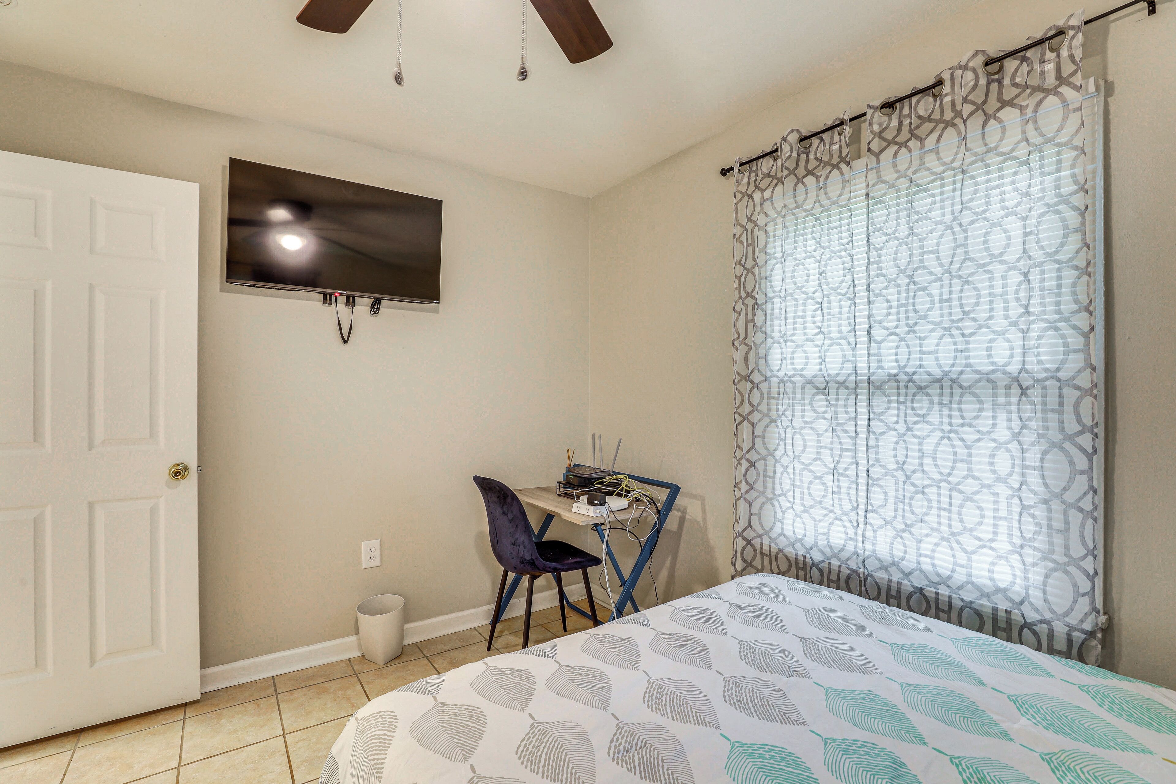 A bedroom with a bed, a desk with a chair, a television on the wall, and a window with patterned curtains.
