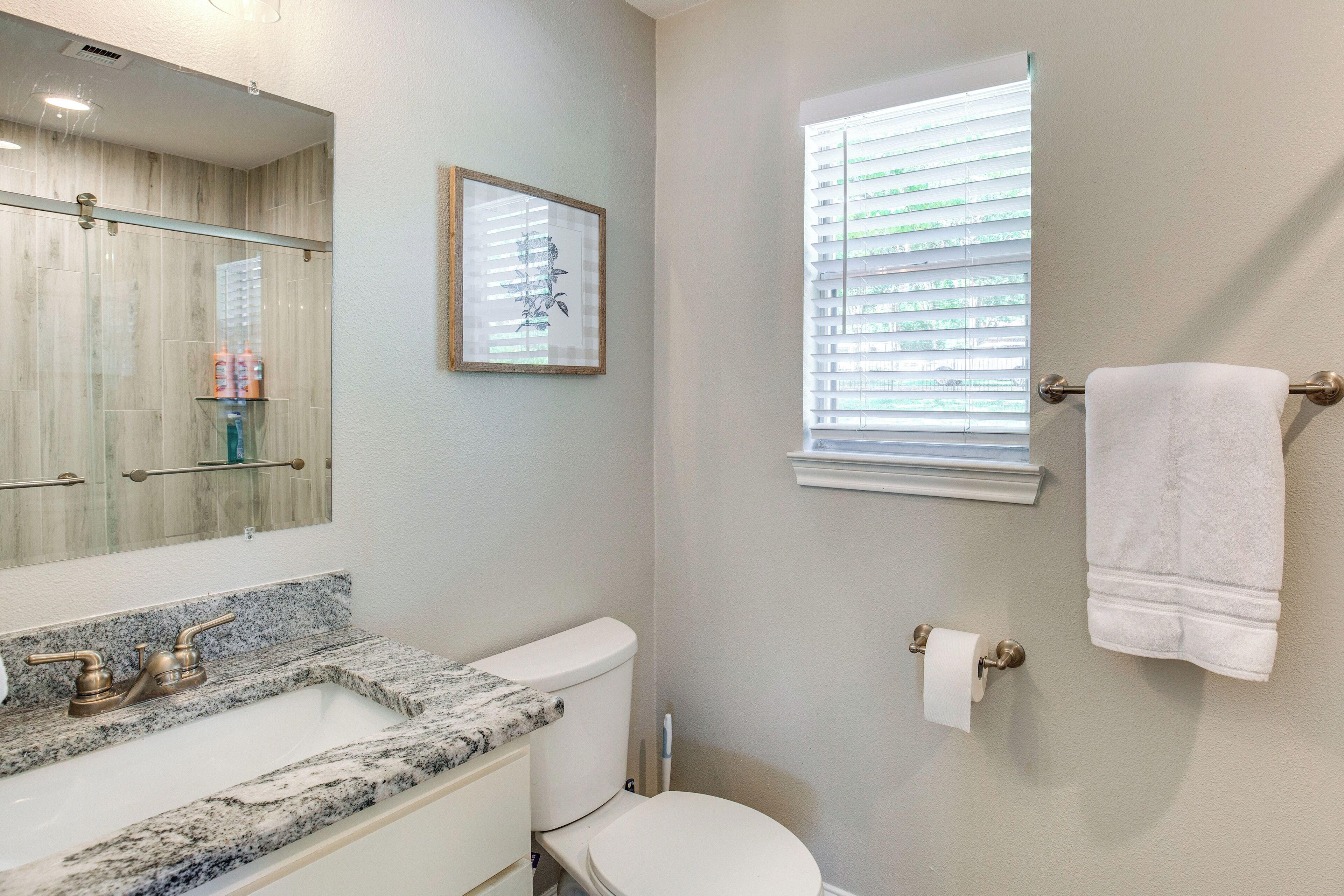 Une salle de bain avec un lavabo en marbre, des toilettes, une œuvre d’art encadrée et une fenêtre avec des stores.
