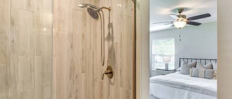 A modern bathroom with a walk-in shower, a ceiling fan, and a bed with a white bedspread and pillows in the adjacent bedroom.
