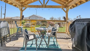 A covered outdoor patio with wooden beams and string lights, featuring a table and chairs, a covered grill, and a view of houses in the background.