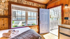 Une pièce avec un lit, une fenêtre donnant sur un lac, une porte menant à l’extérieur et un mur en pierre.
