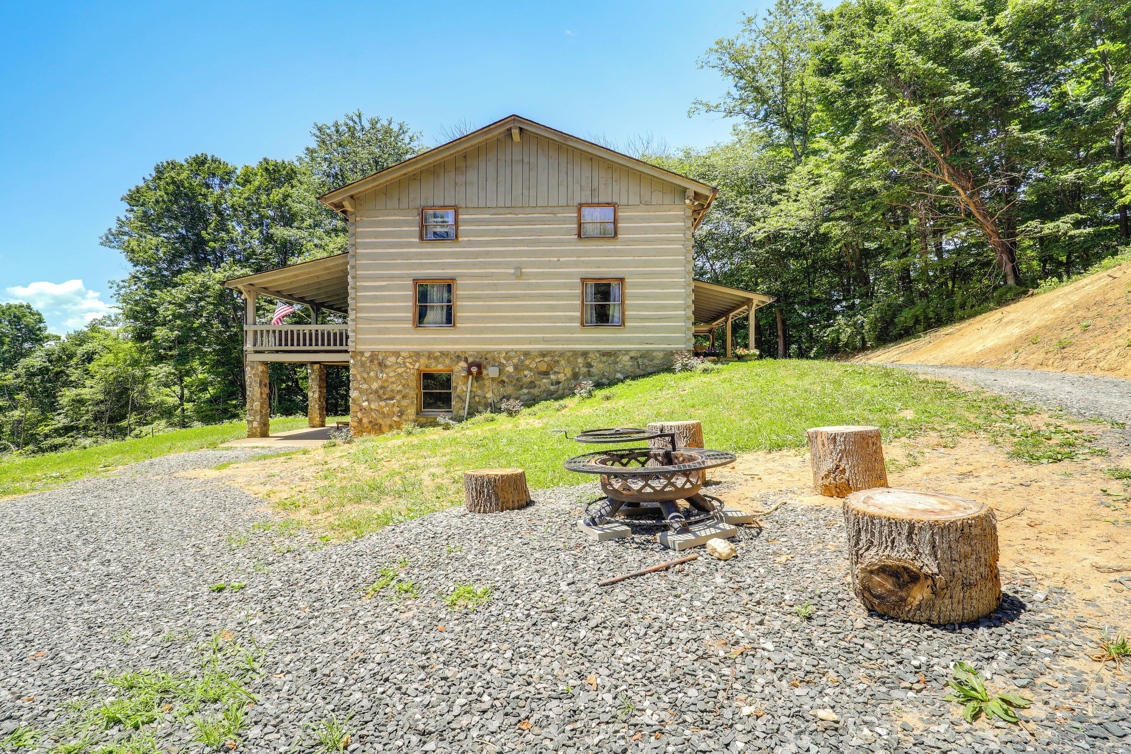 A two-story log cabin with a porch, surrounded by a gravel driveway and a forested hillside.