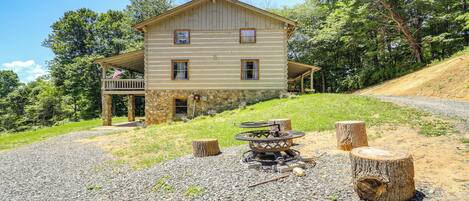 A two-story log cabin with a porch, surrounded by a gravel driveway and a forested hillside.