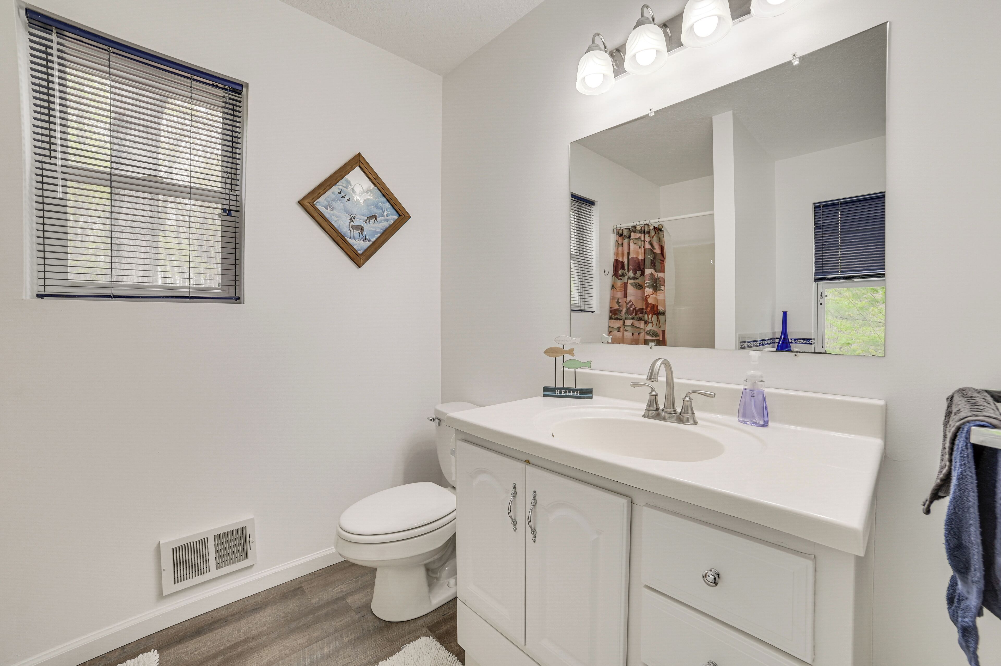 A bathroom with a white sink, mirror, and toilet. There is a window with blinds, a vent, and a framed picture on the wall.
