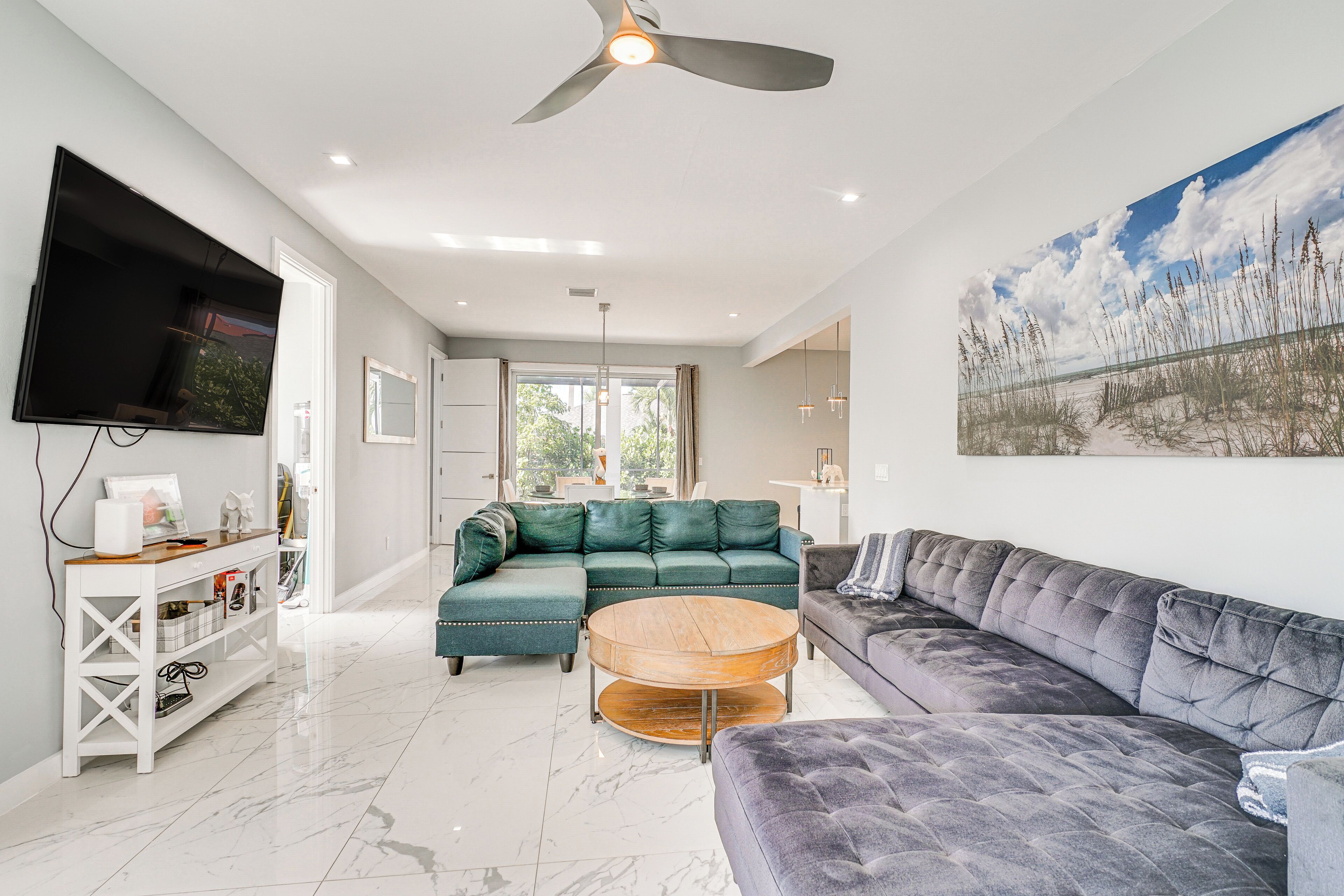 A spacious living room with a large flat-screen TV, a ceiling fan, and a large window offering a view of the outdoors.