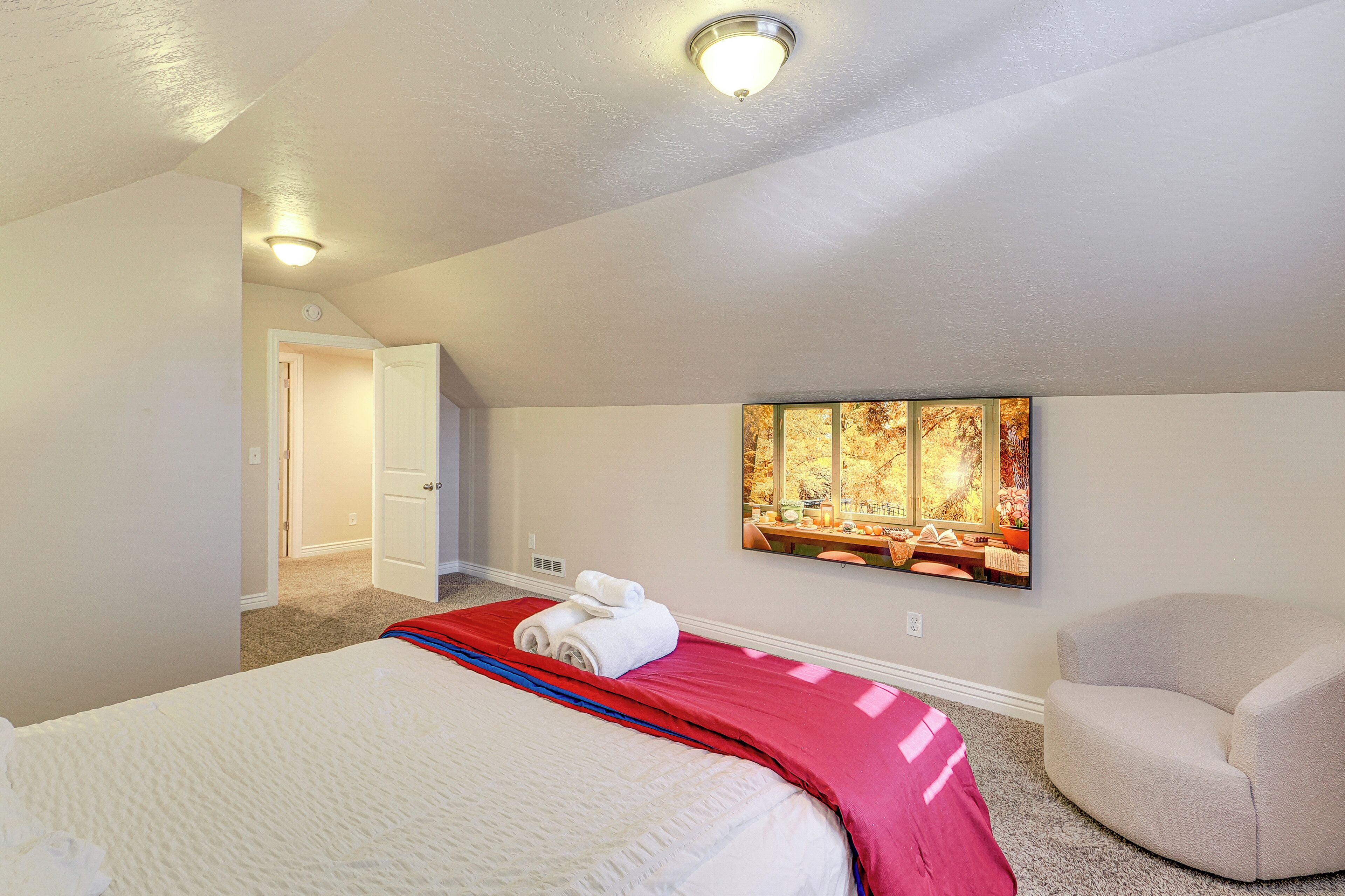 A hotel room with a bed, a red blanket, a beige armchair, and a painting on the wall.