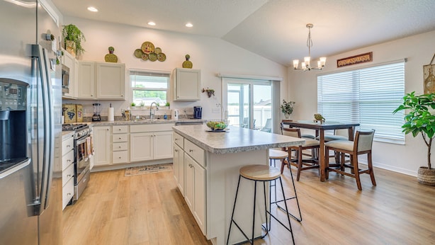 A spacious kitchen with white cabinets, a stainless steel refrigerator, and a wooden dining table with bar stools. - Furnished Lanai & Golf Cart: the Villages Home! (Wildwood)