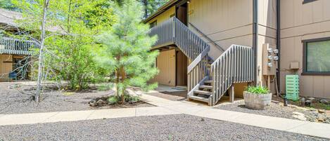 A building with a wooden exterior, a staircase leading to the entrance, and a small garden area with a tree and a planter.