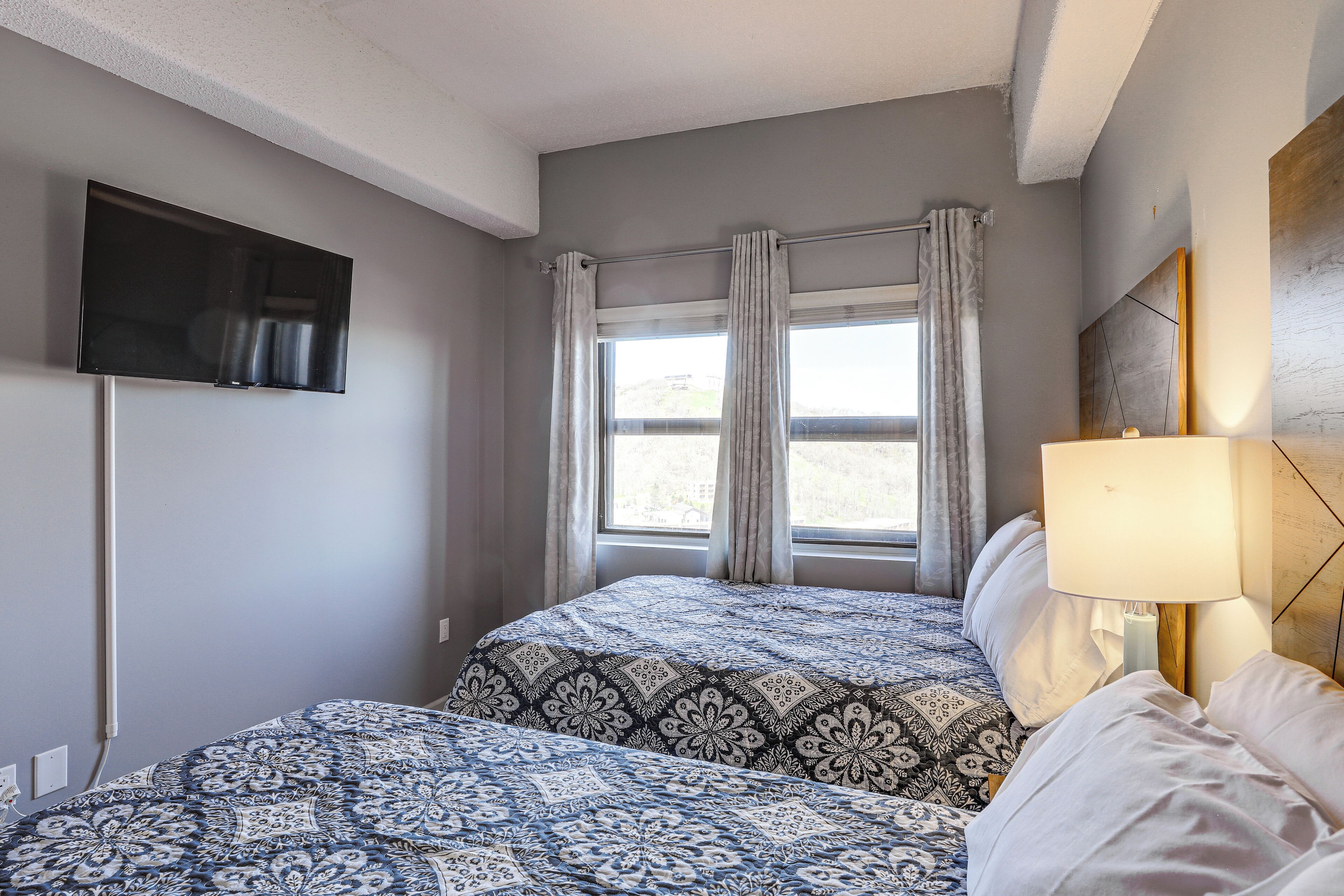A hotel room with two beds, a TV mounted on the wall, a window with curtains, and a bedside lamp.