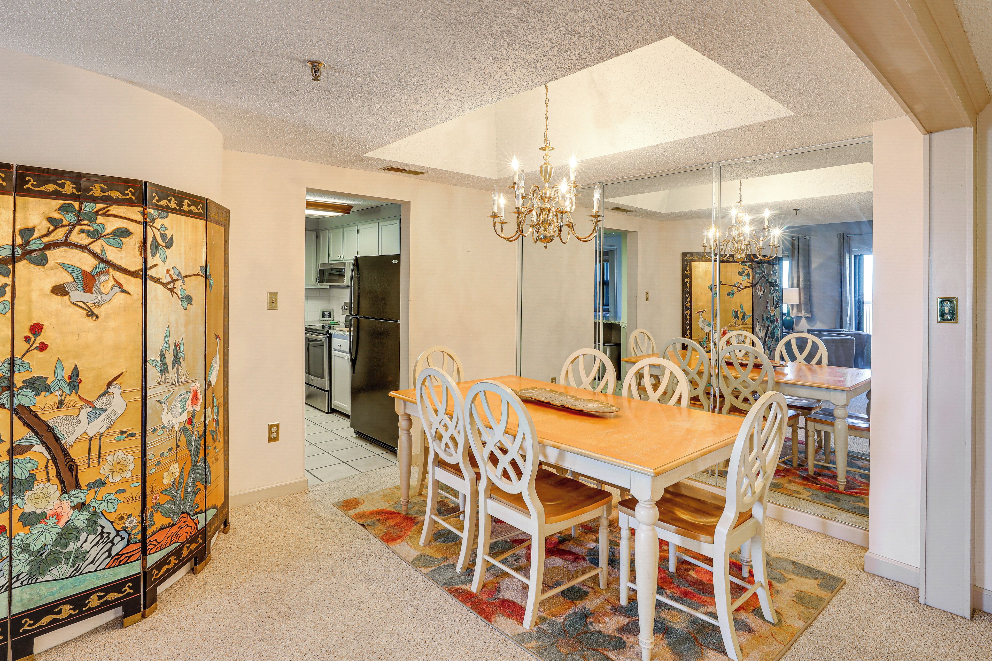 A dining area with a wooden table and white chairs, a chandelier, and a decorative screen with a nature scene.