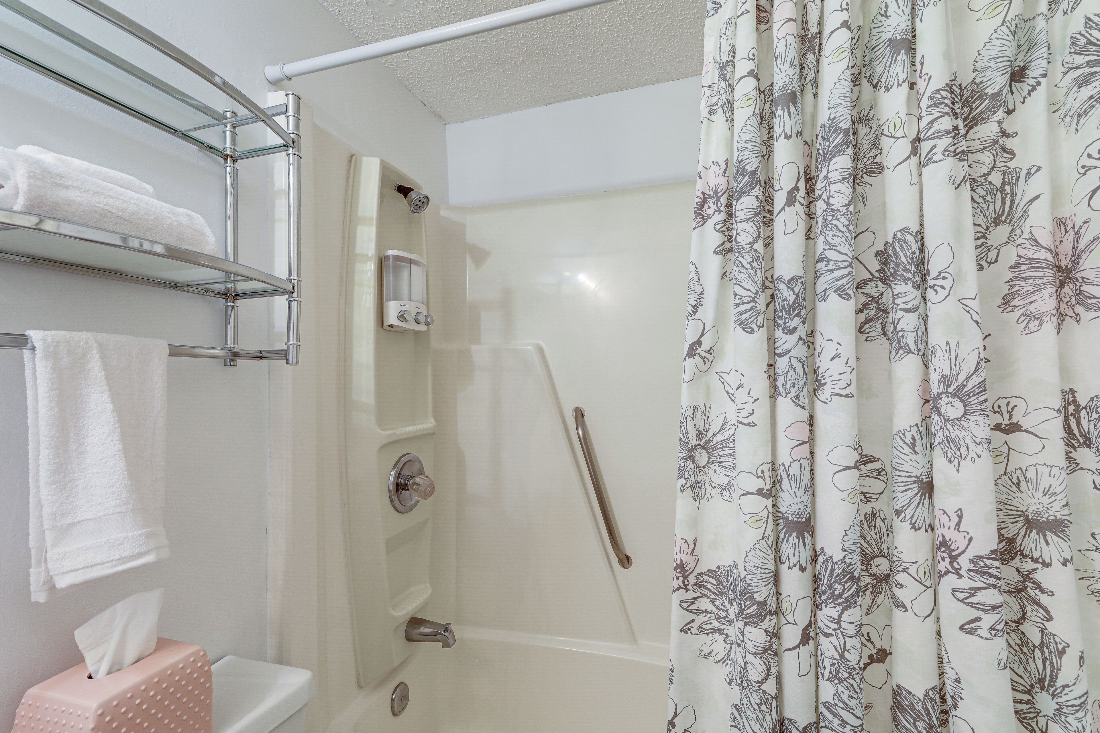 A bathroom with a glass shower enclosure, a towel rack with folded towels, a soap dispenser, and a floral curtain.