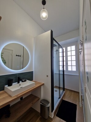 A modern bathroom with a wooden vanity, a round mirror with a light, a glass shower enclosure, and a black mat at the entrance.