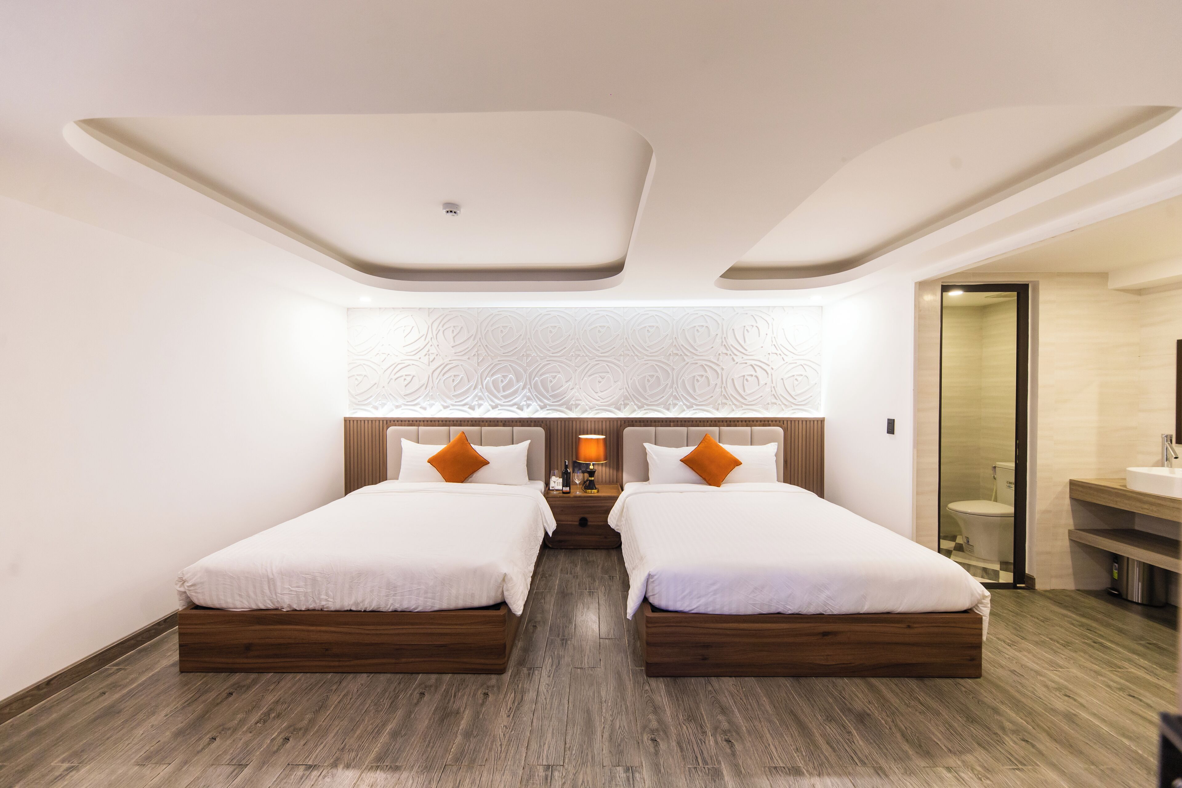 A modern hotel room with two beds, a wooden floor, a patterned wall, and a bathroom visible through a glass door.