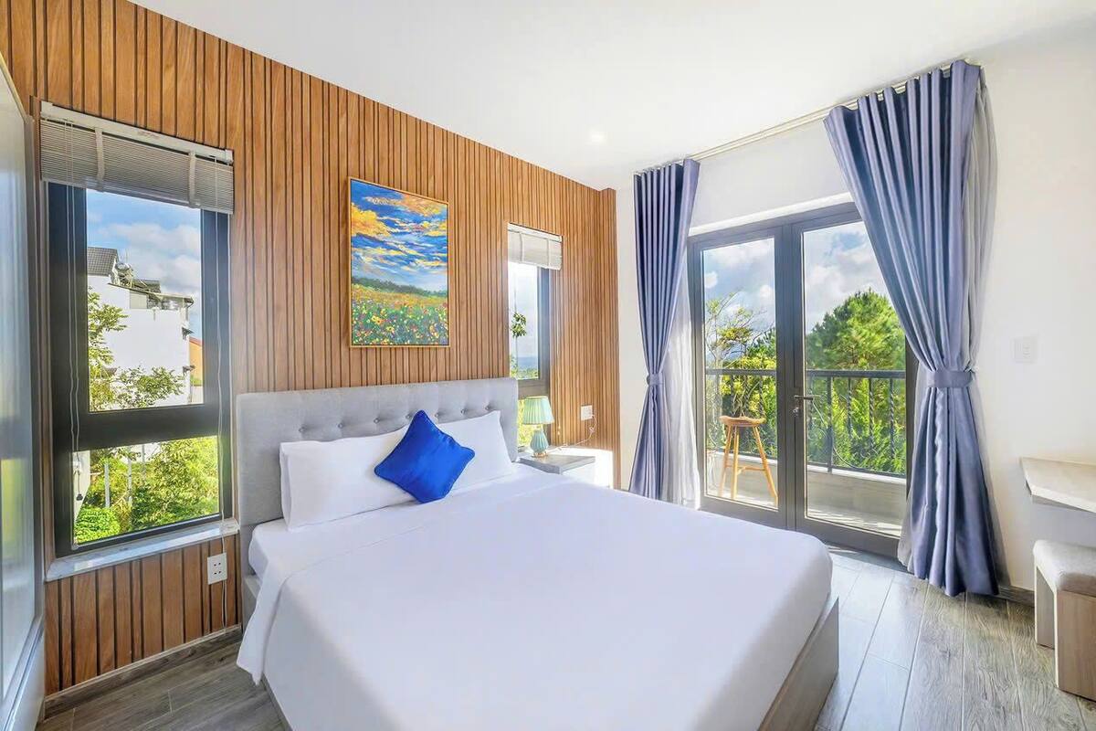 A modern hotel room with a large bed, wooden paneling, a painting, and a balcony with curtains.