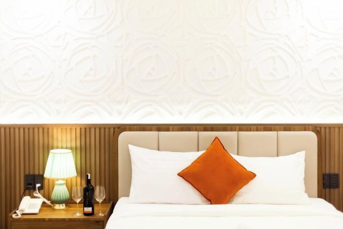 A hotel room with a wooden headboard, a bedside lamp, a phone, a bottle, and two wine glasses on the nightstand.