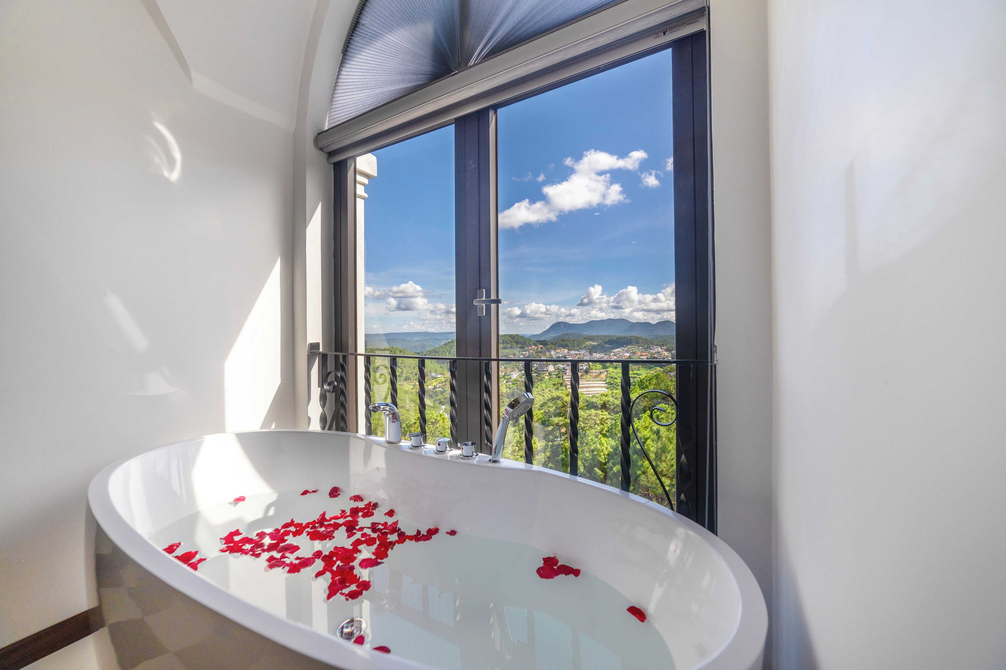 A bathroom with a bathtub filled with rose petals, a large window with a view of greenery and mountains, and a balcony with a railing.