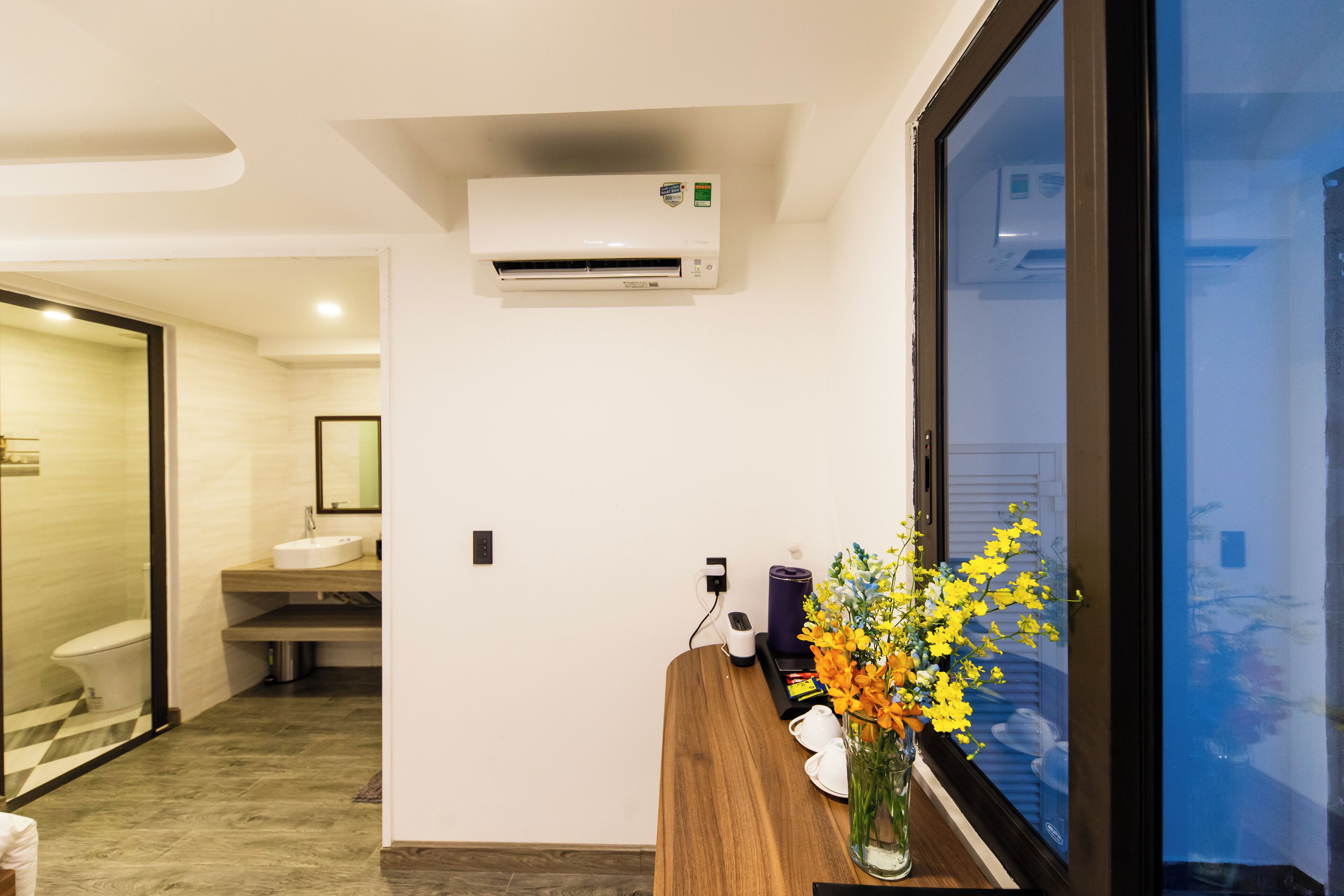 Une salle de bain moderne comprenant des toilettes, un lavabo et un espace douche. On y trouve un meuble-lavabo en bois orné d'un vase de fleurs et une fenêtre offrant une vue agréable.