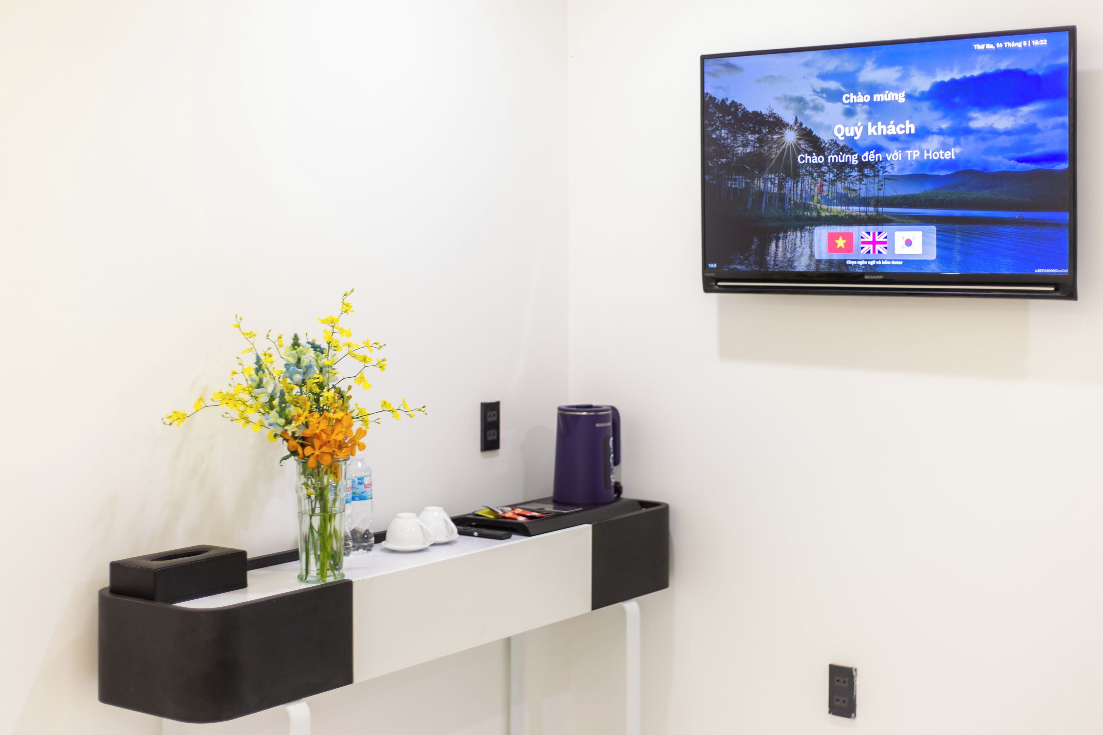 Un mostrador de recepción de hotel con un televisor de pantalla plana mostrando un paisaje escénico, un florero con flores amarillas y una máquina de café.
