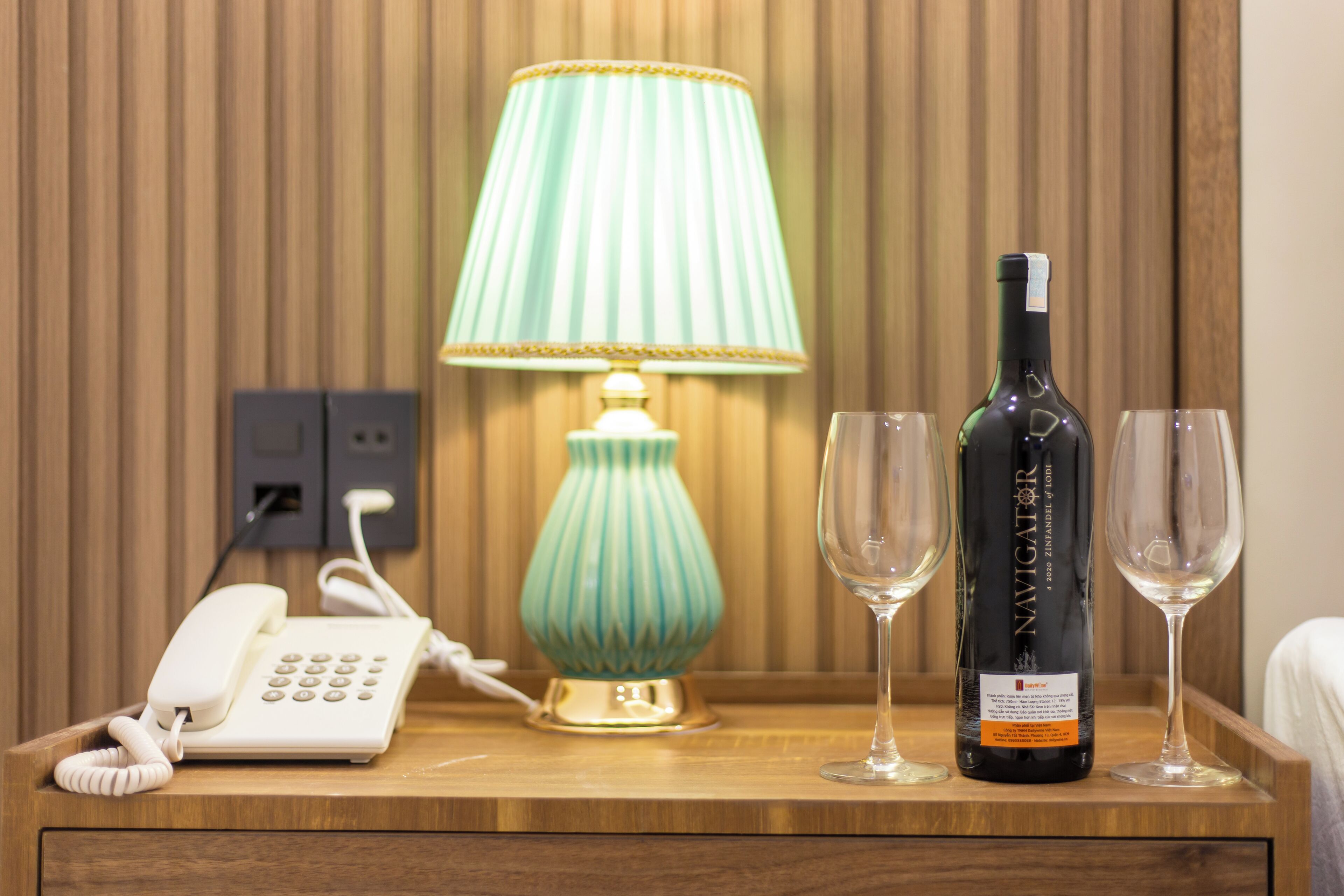 A wooden bedside table with a white telephone, a green lamp, a bottle of wine, and two wine glasses.