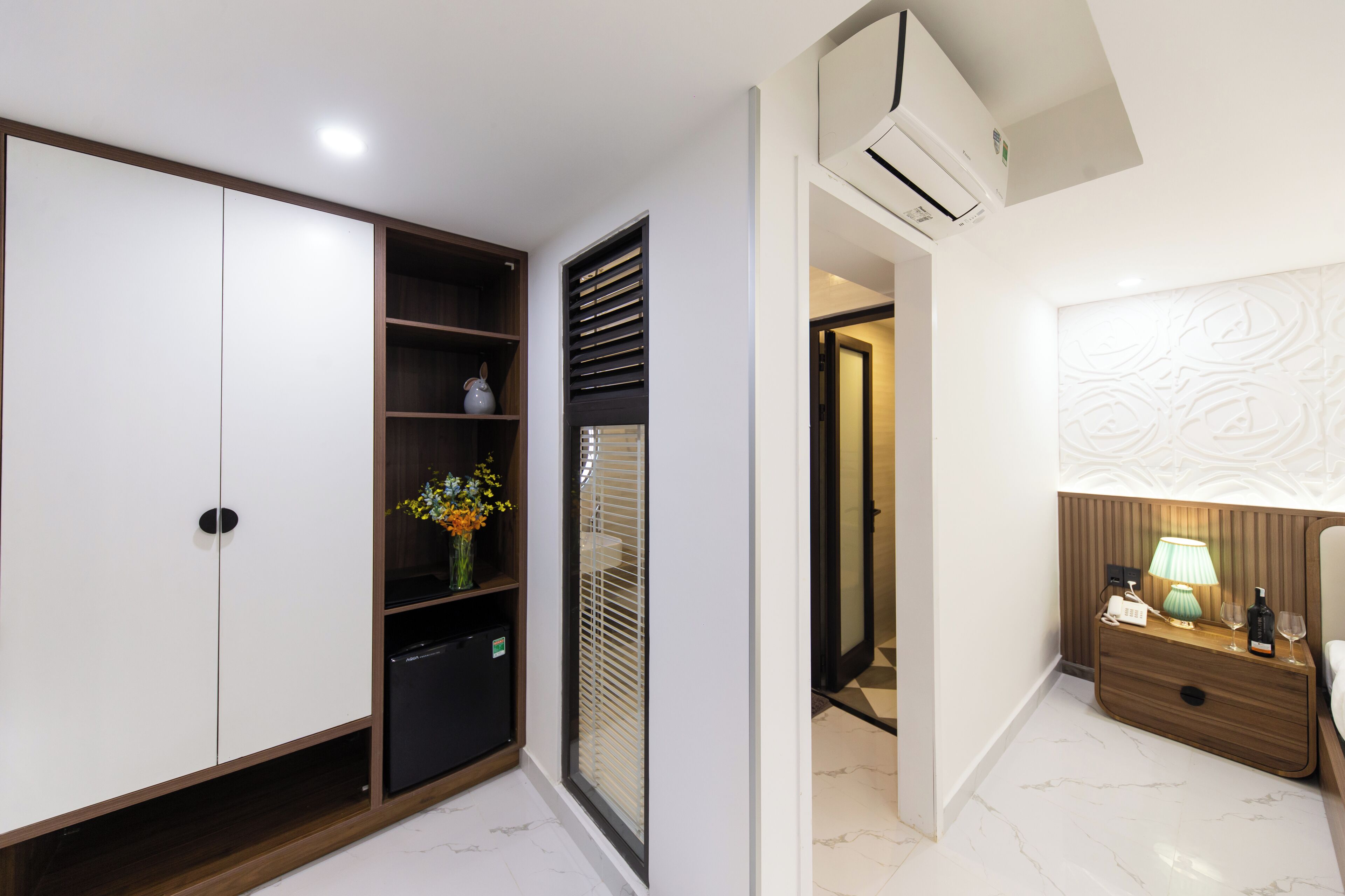 A modern hotel room with a wardrobe, a bedside table with a lamp, and a decorative wall panel.