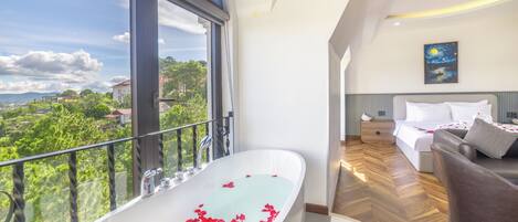 A modern bathroom with a freestanding bathtub filled with rose petals, a large window offering a scenic view, and a bedroom with a bed and a nightstand.