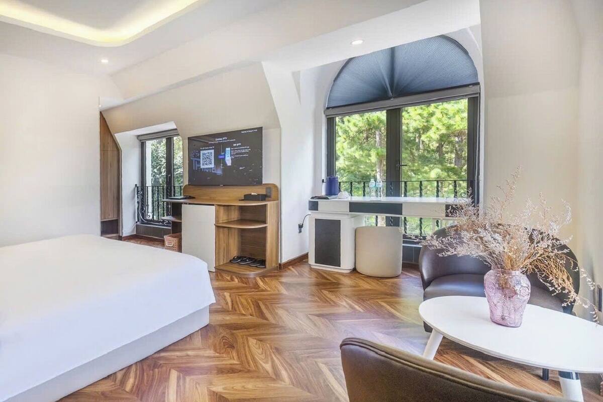 A modern hotel room with a large bed, a dining area with a table and chairs, a sofa, a TV, and a large window offering a view of trees.