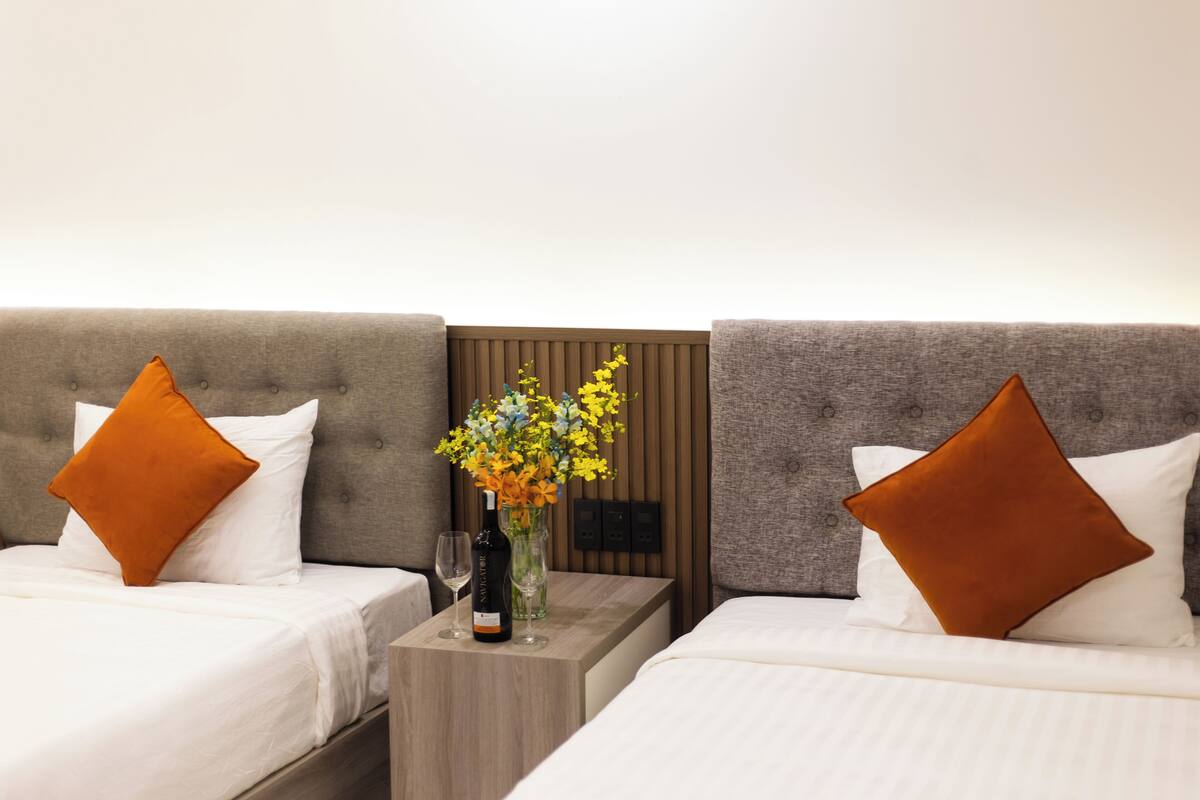 A hotel room with two beds, each with a brown pillow and white bedding. A small wooden nightstand holds a wine bottle and a vase with flowers.