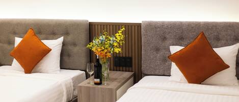 A hotel room with two beds, each with a brown pillow and white bedding. A small wooden nightstand holds a wine bottle and a vase with flowers.