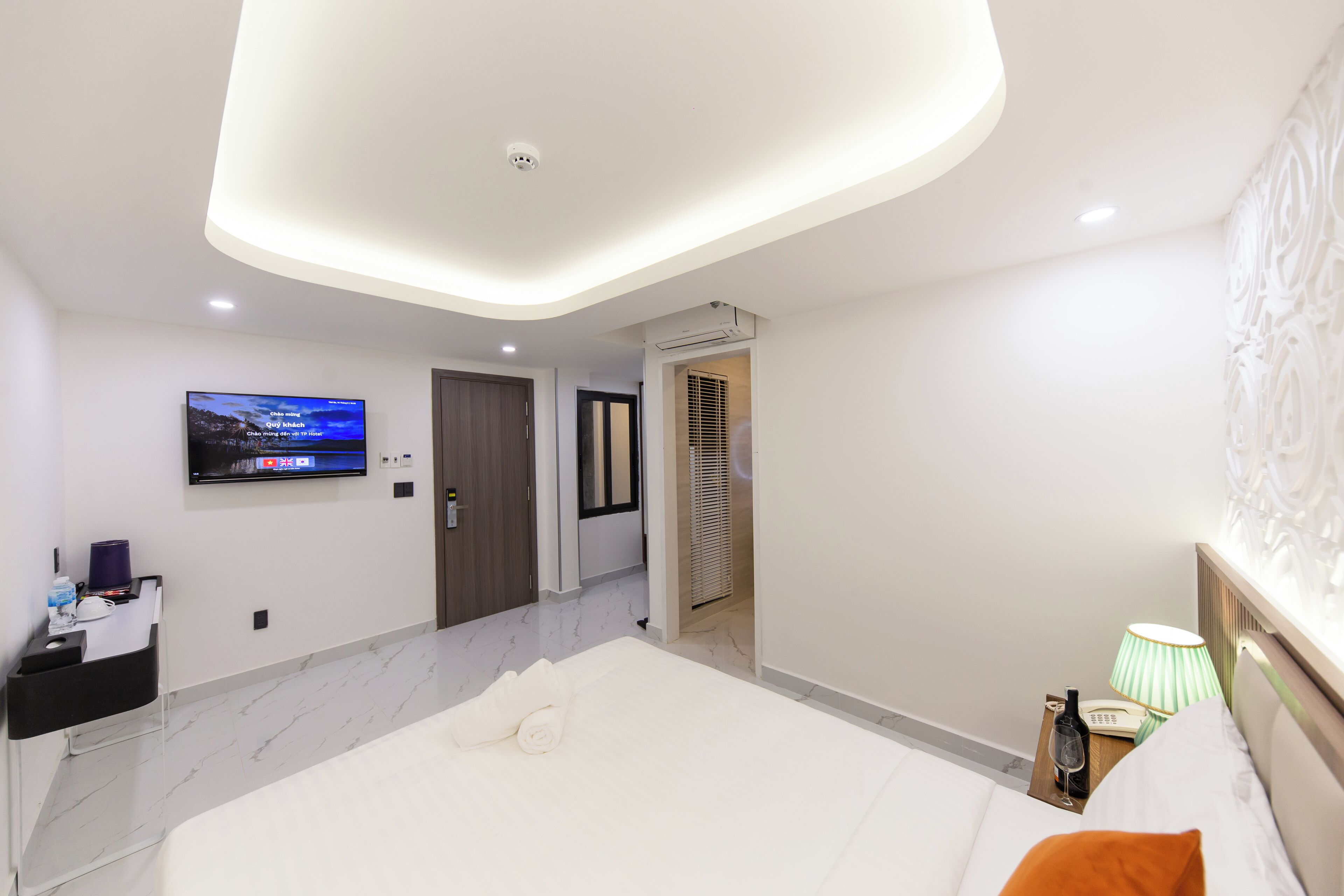 A modern hotel room with a large bed, a flat-screen TV mounted on the wall, a bedside table with a lamp, and a door leading to another room.