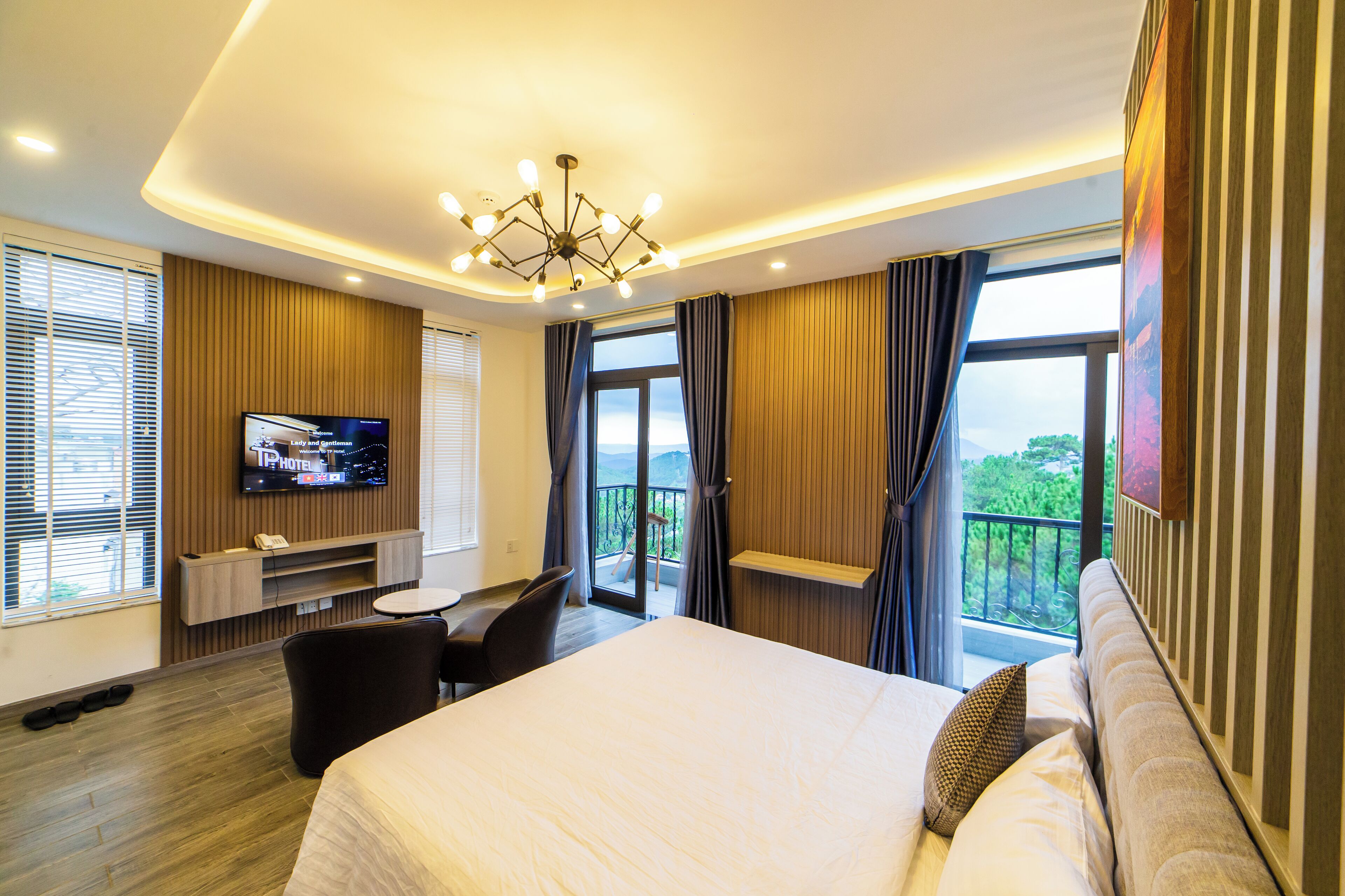 A modern hotel room with a large bed, a flat-screen TV mounted on the wall, a desk with a chair, and a balcony with a view of the outdoors.