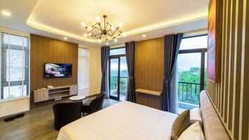 A modern hotel room with a large bed, a flat-screen TV mounted on the wall, a desk with a chair, and a balcony with a view of the outdoors.