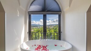 A bathroom with a large bathtub filled with rose petals, a window with a view of a cityscape and mountains, and a balcony railing. - TP Dalat Hotel (Xuan Huong - Da Lat)