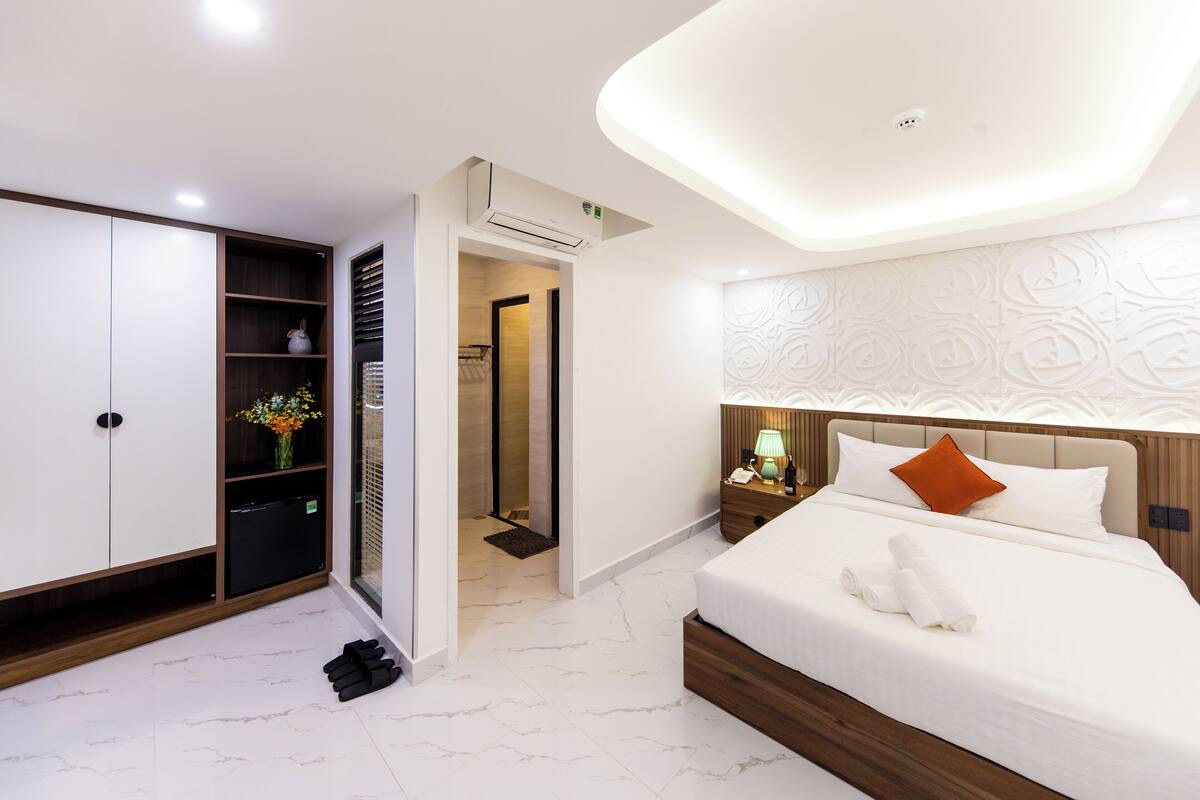A modern hotel room with a neatly made bed, a bedside table with a lamp, a wardrobe, and an air conditioning unit.
