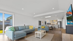 Un salon moderne avec un canapé d’angle, une table basse et un téléviseur. On y trouve également un coin repas avec une table et des chaises, et une cuisine à l’arrière-plan.