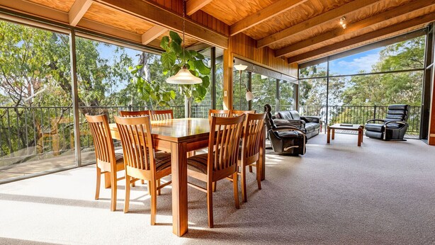 A spacious room with a wooden dining table and chairs, a leather sofa, and a large window offering a view of trees. - Anchorage Views: Indulge in the ultimate coastal getaway at Anchorage Views (Metung)